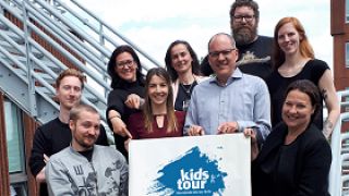 astendo unterstützt 28. Internationale kids-tour Berlin astendo-team haelt plakat mit logo kids-tour berlin in die Kamera