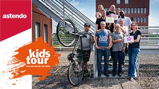 astendo-team unterstuetzt-internationale-kids-tour Team von astendo steht auf Treppe, hält Schilder mit "kids-Tour" hoch, daneben ein Kollege mit Fahrradhelm und Fahrrad